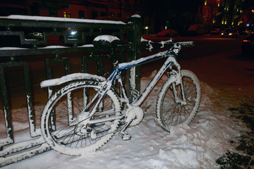 Fototapeta na wymiar A snow-covered bike parked on a fence on a winter street.