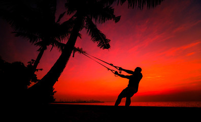 The athlete performs exercises on the beach with fitness straps which are fixed on the palm tree. Sunset
