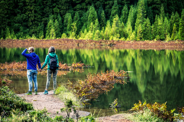 Young couple in colorful sweatshirts is standing in front of the volcanic lake on Azores Islands. Beautiful reflections on the background and beautiful green forest. Portugal. - 247117402