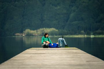 Young just married couple is sitting on pier by the volcanic lake on Azores islands. Low angle view, front view, soft focus. Mountain on background. - 247117453
