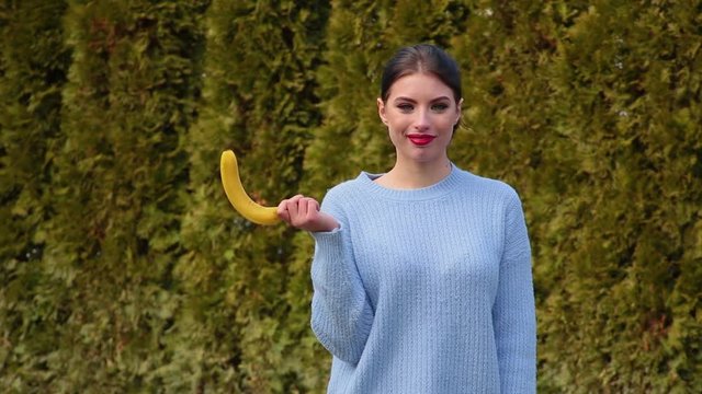 Portrait attractive young woman in blue jacket holds banana in her hand then lowers it and lifts it up imitating male erection, talking oops and smiling against green trees