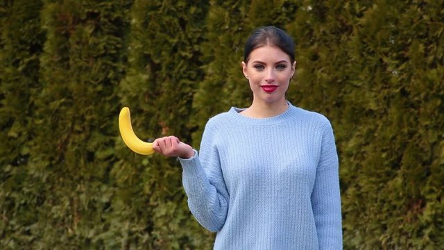Portrait attractive young woman in blue jacket holds banana in her hand then lowers it and lifts it up imitating male erection, talking oops and smiling against green trees