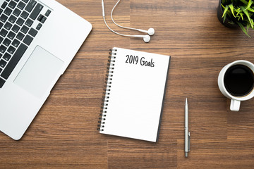 Notebook with 2019 Goals text is on top of wood office desk table with supplies. Top view with copy space, flat lay.