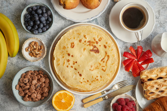 Breakfast table setting with fresh fruits, pancakes, coffee, croissants, sweet cakes, muffins. Flat lay on stone table.