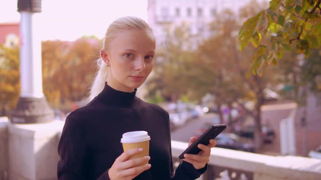 girl using smartphone texting message holding coffee to go looking at the camera standing outdoors. young caucasian woman with blond hair posing in autumn city.