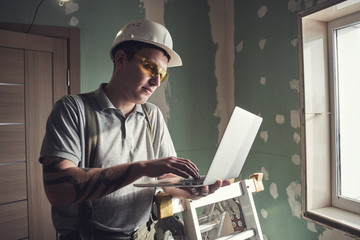 Room repair. Builder in helmet and glasses stands with a laptop in his hands on the background of the construction