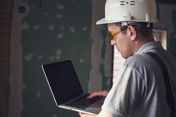 Room repair. Builder in helmet and glasses stands with a laptop in his hands on the background of the construction