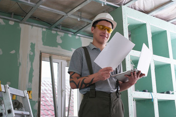 Room repair. Builder in helmet and glasses stands with a laptop and drawings in his hands on the background of the construction