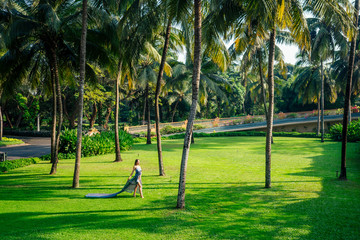fashion outdoor photo of beautiful young woman with long hair in luxurious gray silk dress posing in a tropical forest with palm trees and flowers
