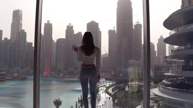 Young woman coming to the window and looking and looking down to the lake and skyscrapers at the subset.