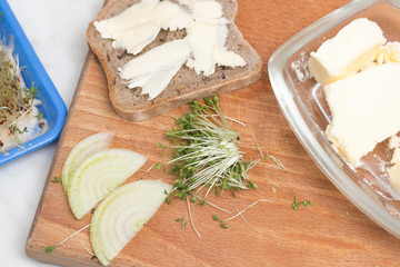 Obcięta porcja rzeżuchy leży na desce do krojenia w towarzystwie kromki chleba posmarowanej masłem. Masło w maselniczce, cebula leży obok.