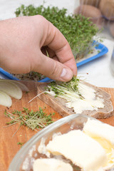 Męska dłoń układa obcięte kiełki rzeżuchy na kromce chleba posmarowanej masłem. 