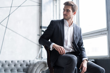 Portrait of confidence. Thoughtful young man in full suit looking away while sitting on the stool.