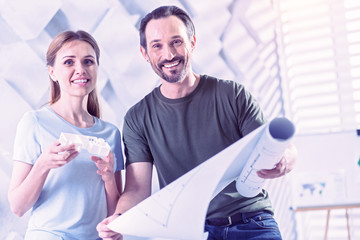 Happy architect holding blueprint while his colleague standing with house miniature