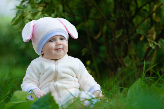 Baby girl with chubby cheeks in rabbit costume seating on grass in park.