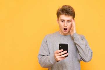 Portrait of a shocked guy standing with a smartphone in his hand on a yellow background, looking at the screen, took a hand on his head against the yellow background. Copyspace