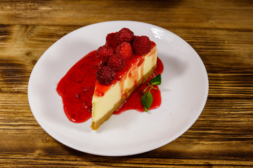 Piece of tasty New York cheesecake with raspberries and raspberry jam in a white plate on wooden table
