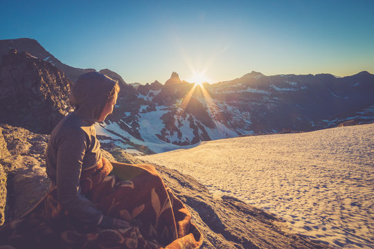 Girl sitting on a rock and covered with a blacket is watching Sunset From the top of the Mountain in a glacier area in the Alps with snow and blue sky