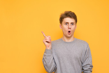 Shocked guy on a yellow background looks at the camera and shows his finger on the blank space for...