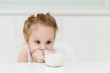 Little smart caucasian girl drinks milk from a glass cup in the kitchen and has fun, healthy eating concept for children.