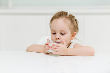 Little smart caucasian girl drinks milk from a glass cup in the kitchen and has fun, healthy eating concept for children.