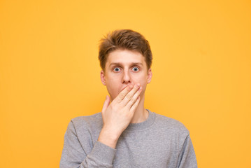 Close-up portrait of a surprised boy that covers his mouth with his hand looks emotionally into the camera on a yellow background.