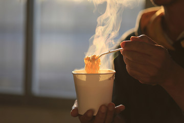 Man hand holding plastic fork of instant noodles with smoke rising in the home sunset background,...