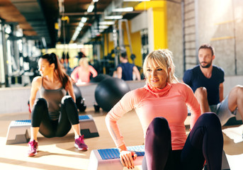 Group of dedicated people doing exercises on stepper in gym. In background their reflection in...