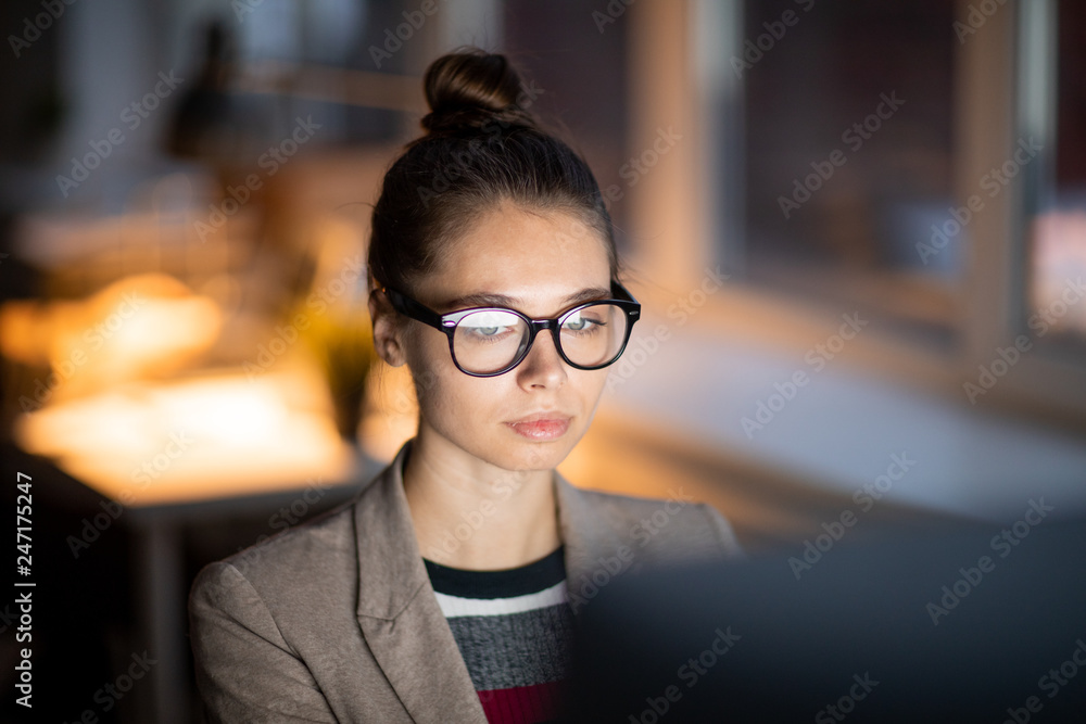 Poster young casual businesswoman in eyeglasses concentrating on working in front of computer monitor in of
