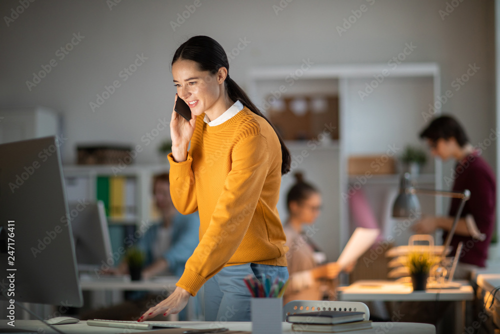 Fototapete young casual manager standing by desk in front of computer monitor, looking at its screen and talkin