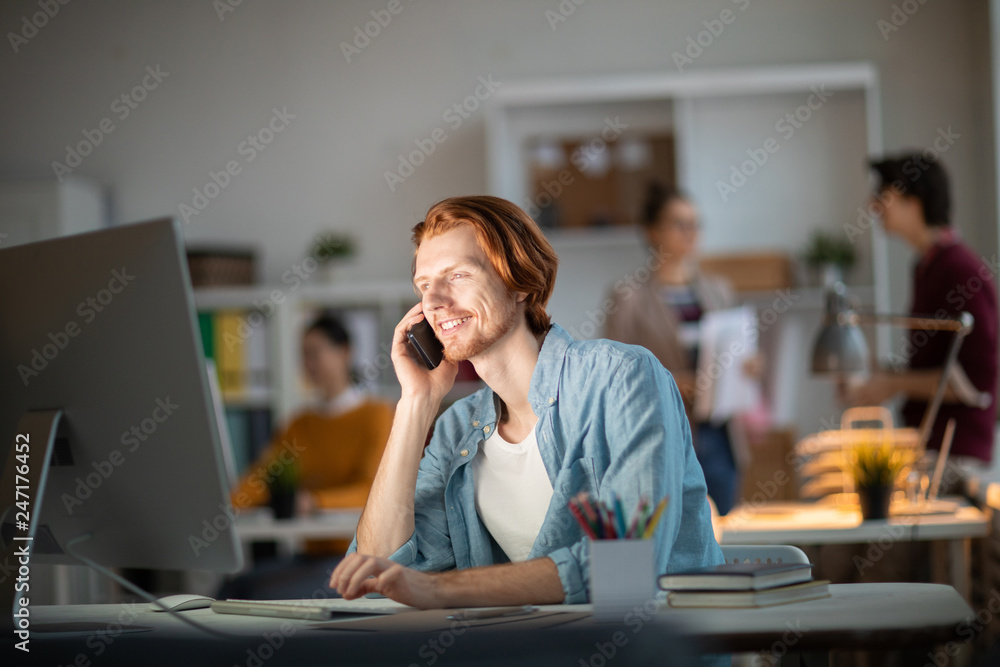 Poster happy young businessman with smartphone by his ear looking at computer screen while working with web