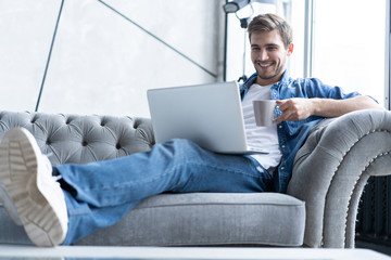 Young attractive smiling guy is browsing at his laptop, sitting at home on the gray sofa at home, wearing casual outfit.