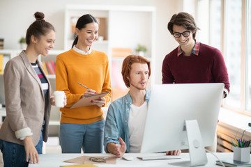 Several happy young developers standing by desk in front of computer monitor and making notes while...