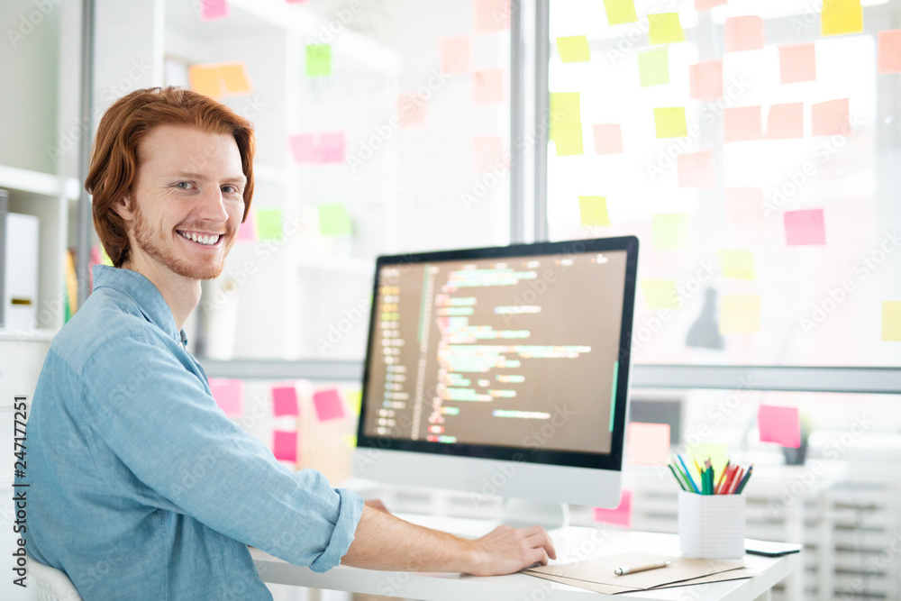 Wall mural young cheerful it-developer with toothy smile looking at you while sitting by workplace in front of 