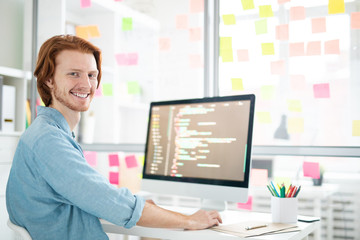 Young cheerful it-developer with toothy smile looking at you while sitting by workplace in front of...