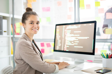 Young casual woman sitting by desk in front of camera and analyzing coded data on computer screen