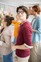 Obraz premium Young smiling handsome manager in casualwear looking at you in office environment with working colleagues