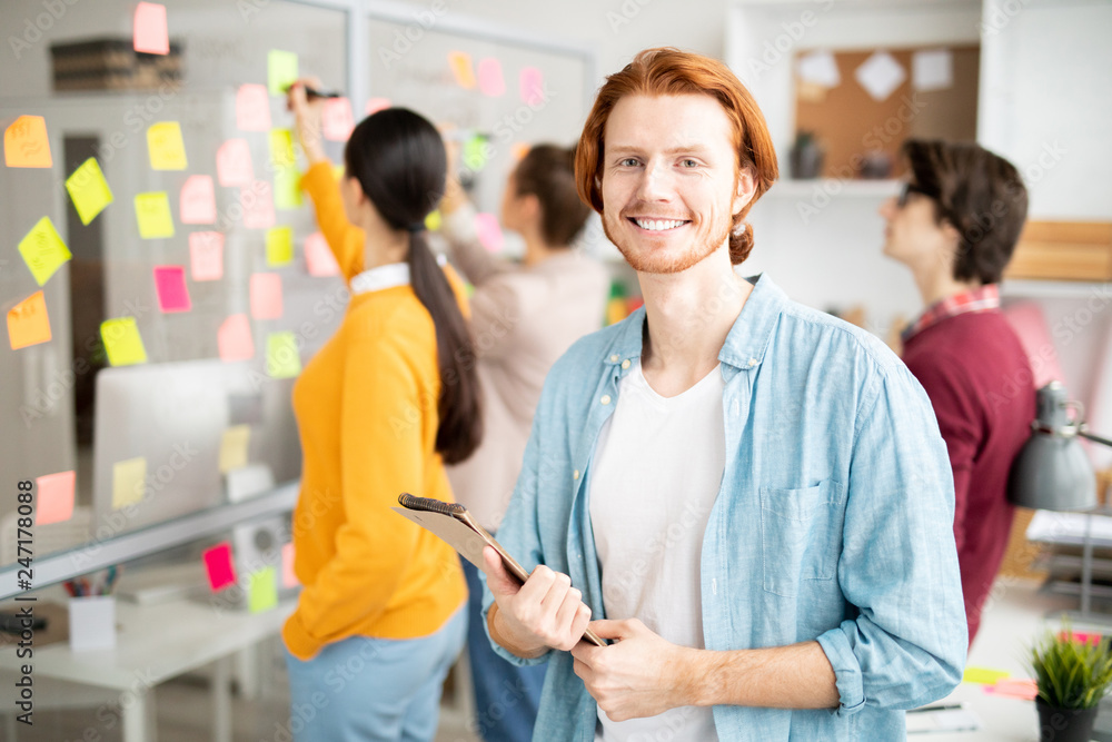 Wall mural happy young smiling businessman with notepad standing in front of camera in working environment