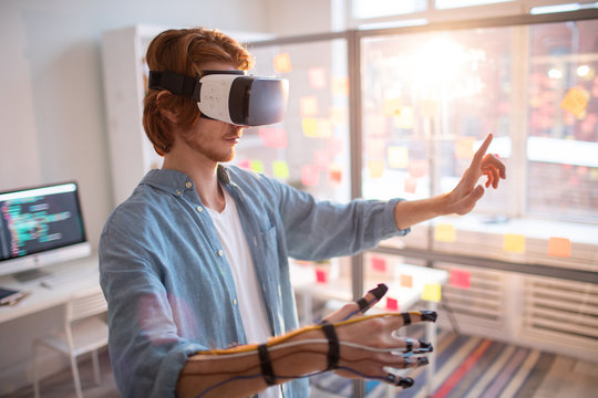 Young contemporary businessman in vr headset with sensors on fingers touching virtual display while making presentation