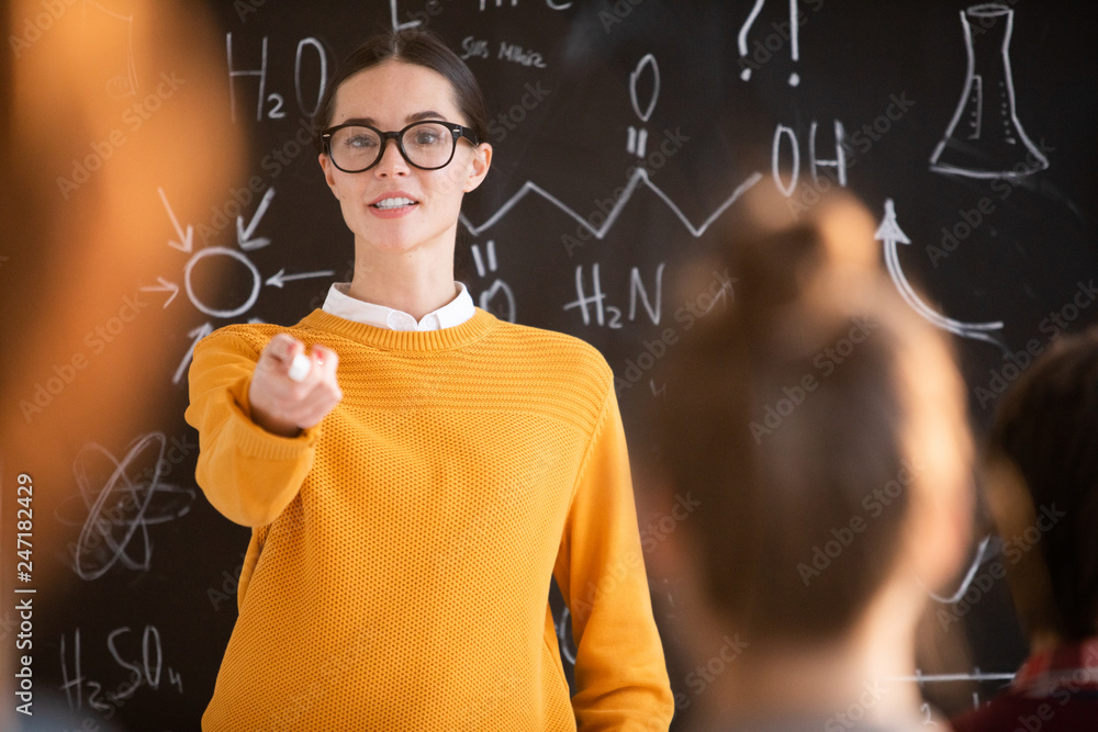 Poster Young teacher in casual pullover and eyeglasses pointing at one of students during explanation of new topic at lesson