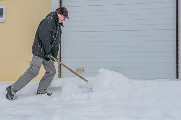 A man in a black jacket and light-colored gray pants and a brown hat cleans snow with a wooden shovel in winter