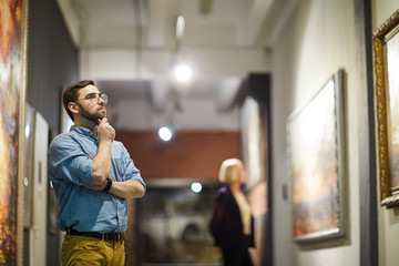 Portrait of pensive bearded man looking at paintings standing in art gallery or museum, copy space