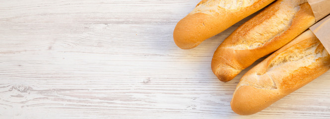 French baguettes in paper bags on white wooden background, overhead view. Space for text.