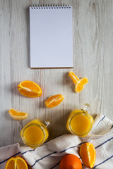 Fresh orange juice in glass jars, blank notebook over white wooden surface, top view. Flat lay, from above, overhead.
