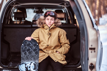A teenage snowboarder dressed in warm clothes sitting on the trunk of a car. Outdoor activities...