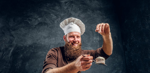 Cheerful bearded baker sprinkling some flour on his freshly made muffin next to a table in a dark...