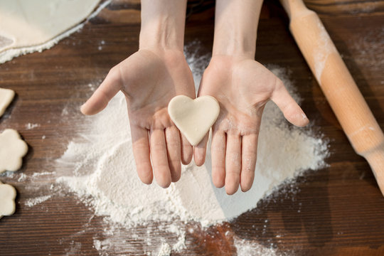 Overview of raw dough in shape of heart on female hands over pile of sifted flour during cooking