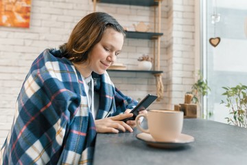 Autumn winter portrait of smiling teen girl with mobile phone and cup of coffee in coffee shop, girl covered with woolen plaid blanket