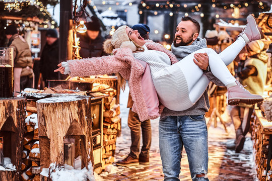 An attractive couple in love. A handsome man holding his girlfriend on hands, having fun together while standing at the winter fair at a Christmas time