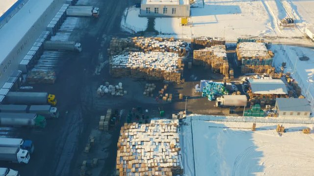 Aerial look-down shot of a storage yard of wooden pallets with working forklifts on the territory of a logistics hub/ warehouse with ramps and semi-trailers trucks stands on load/ unload goods